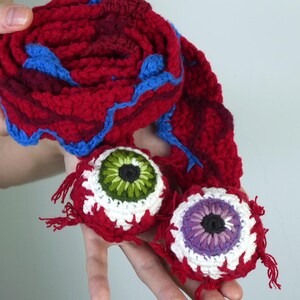K&ouml;nnte beinhalten: Handgefertigtes H&auml;kel-Set mit einem rot-blauen Schal und zwei augenf&ouml;rmigen Ornamenten. Die Augen haben wei&szlig;e, rote, gr&uuml;ne und violette Details. Der Schal ist aufgerollt und hat blaue Verzierungen.