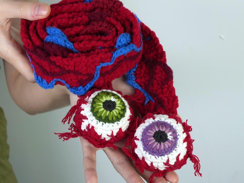 K&ouml;nnte beinhalten: Handgefertigtes H&auml;kel-Set mit einem rot-blauen Schal und zwei augenf&ouml;rmigen Ornamenten. Die Augen haben wei&szlig;e, rote, gr&uuml;ne und violette Details. Der Schal ist aufgerollt und hat blaue Verzierungen.