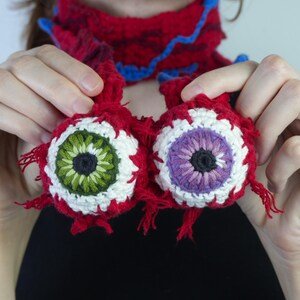 K&ouml;nnte beinhalten: Zwei geh&auml;kelte, augenf&ouml;rmige Ornamente mit roten, wei&szlig;en, gr&uuml;nen und violetten Details. Die Augen werden vor einem roten und blauen Schal gehalten. Die handgefertigten Ornamente haben ein einzigartiges, skurriles Design, ideal f&uuml;r Halloween.