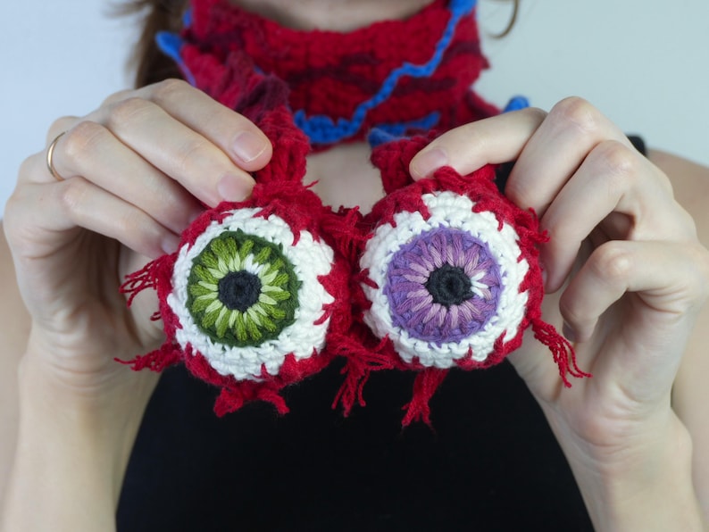 K&ouml;nnte beinhalten: Zwei geh&auml;kelte, augenf&ouml;rmige Ornamente mit roten, wei&szlig;en, gr&uuml;nen und violetten Details. Die Augen werden vor einem roten und blauen Schal gehalten. Die handgefertigten Ornamente haben ein einzigartiges, skurriles Design, ideal f&uuml;r Halloween.