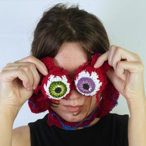 K&ouml;nnte beinhalten: Handgefertigte, geh&auml;kelte Accessoires in Augenform in Rot, Wei&szlig;, Gr&uuml;n und Lila. Die handgefertigten Artikel weisen detaillierte N&auml;hte auf und werden vor dem Gesicht gehalten. Der Hintergrund ist eine schlichte wei&szlig;e Wand.