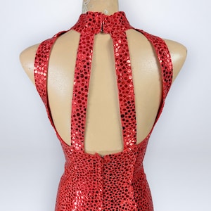 May include: A sleeveless red dress covered in small, reflective circular sequins. The garment features a high mock neckline and a distinctive open-back design with two vertical straps connecting the collar to the bodice, creating a cutout effect.