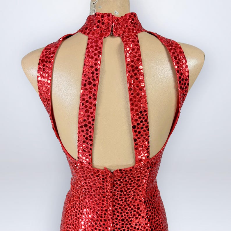 May include: A sleeveless red dress covered in small, reflective circular sequins. The garment features a high mock neckline and a distinctive open-back design with two vertical straps connecting the collar to the bodice, creating a cutout effect.