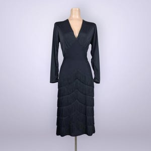 May include: A black, long-sleeved dress with a V-neck and fringe detailing. The dress is displayed on a mannequin and has a fitted bodice that flows into a skirt with layered fringe. The dress is a midi length.