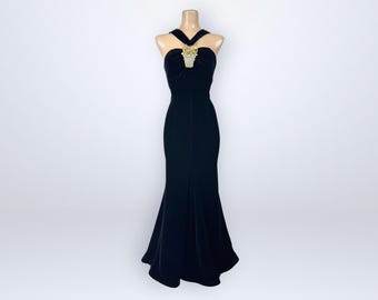 May include: A floor-length black velvet gown featuring a halter neckline with a decorative gold-toned embellishment at the center of the chest. The dress has a fitted bodice that flares slightly at the hem, creating a long, sleek silhouette.