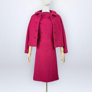 May include: A bright magenta two-piece set consisting of a sleeveless knee-length dress and a matching cropped jacket. The jacket features a collared neckline and a front button closure with dark blue buttons on one side and brown buttons on the other.
