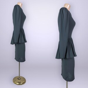 Puede incluir: Un vestido verde oscuro con mangas largas y cintura peplum, exhibido en un maniqu&iacute;. El vestido tiene un corpi&ntilde;o ajustado y una falda hasta la rodilla. El maniqu&iacute; est&aacute; sobre un soporte dorado.