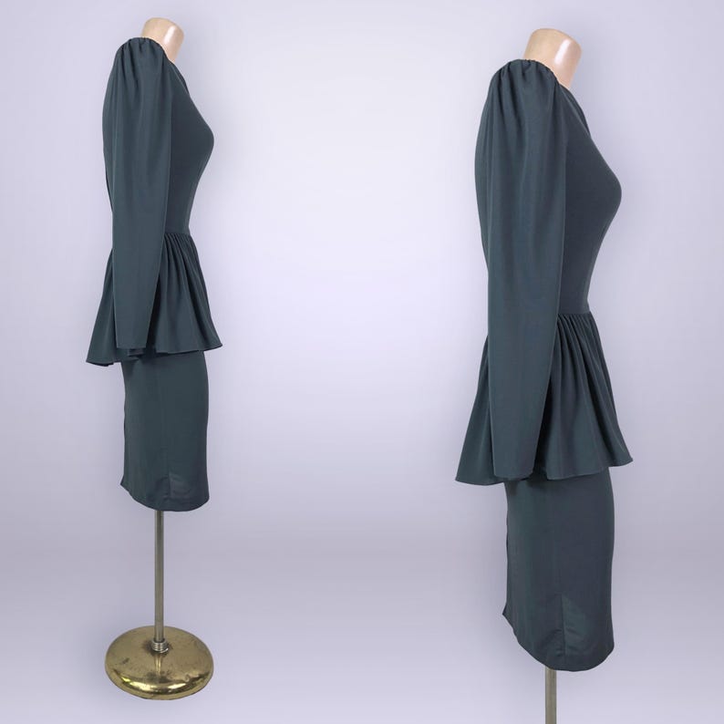 Puede incluir: Un vestido verde oscuro con mangas largas y cintura peplum, exhibido en un maniqu&iacute;. El vestido tiene un corpi&ntilde;o ajustado y una falda hasta la rodilla. El maniqu&iacute; est&aacute; sobre un soporte dorado.