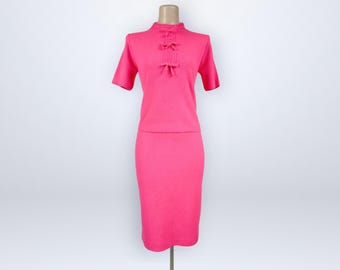 May include: A bright pink two-piece set consisting of a short-sleeved top and a knee-length skirt. The top features a high neckline with a vertical placket adorned with three small decorative bows and a textured knit fabric throughout.
