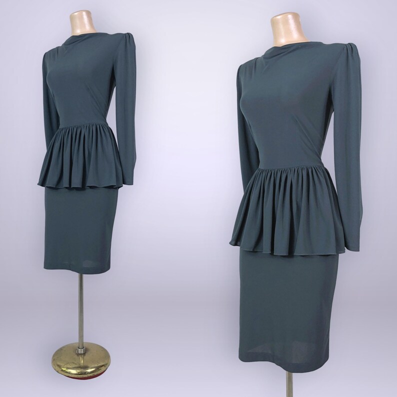 Puede incluir: Dos vestidos de color verde azulado sobre maniqu&iacute;es. Cada vestido tiene mangas largas, cuello alto y cintura peplum. La falda es de estilo l&aacute;piz. Los maniqu&iacute;es est&aacute;n sobre soportes dorados. El fondo es gris claro.