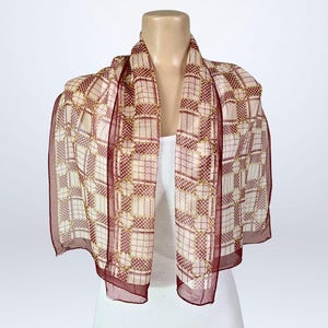Pode incluir: Um lenço transparente com um padrão geométrico em creme, bordô e dourado. O lenço é drapeado sobre um manequim, mostrando sua forma retangular e seu tecido delicado. As bordas são de um bordô mais escuro.