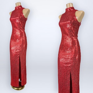 May include: A sleeveless floor-length red dress featuring a high mock neck collar and a repeating black speckled pattern throughout the fabric. The garment includes a straight silhouette with a single vertical slit along the front lower hemline.