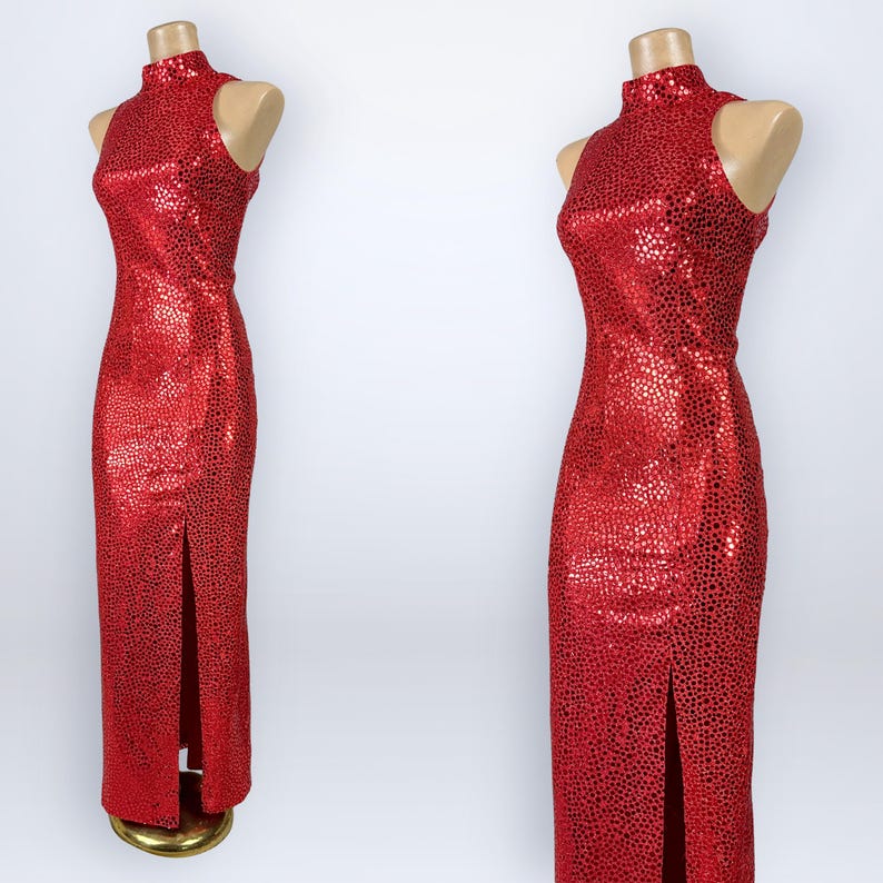 May include: A sleeveless floor-length red dress featuring a high mock neck collar and a repeating black speckled pattern throughout the fabric. The garment includes a straight silhouette with a single vertical slit along the front lower hemline.