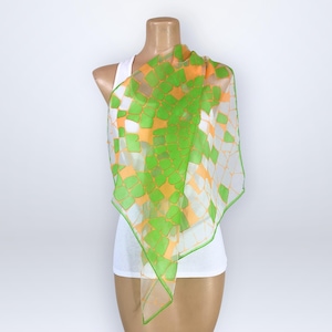 May include: A sheer, square scarf with a geometric pattern of green and orange squares on a white background. The scarf is draped over a mannequin wearing a white tank top. The scarf's edges are outlined in green.