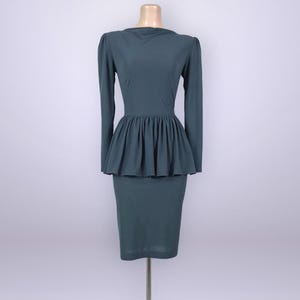 May include: A teal-colored dress with long sleeves and a peplum waist. The dress has a fitted bodice and a knee-length pencil skirt. The dress is displayed on a mannequin against a plain background. The dress is made of a smooth, solid fabric.