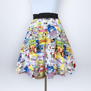 May include: A knee-length skirt featuring a white background with a colorful collage of vintage Disney movie posters. The skirt has a high waist with a black and white polka dot fabric waistband and a flared, A-line silhouette.