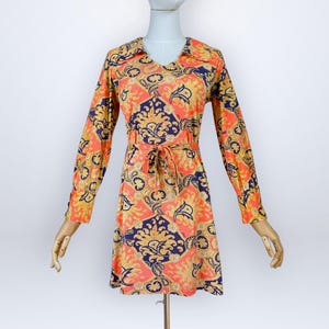 Pode incluir: Um vestido de manga comprida com um padrão vibrante estilo damasco em laranja, dourado e azul-marinho. O vestido inclui um decote em V, um detalhe de colarinho e um cinto de tecido a condizer na cintura. As mangas têm punhos abotoados.