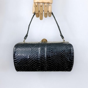 May include: A black handbag with a textured snakeskin pattern, featuring a structured rectangular body, a single top handle, and a gold-toned metal frame closure at the top. The surface has a glossy finish with a distinct scale-like appearance.
