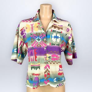 May include: A button-down shirt with a colorful geometric pattern in shades of purple, blue, yellow, and green. The short-sleeved shirt is displayed on a mannequin. The shirt has a collared neckline and button closures.