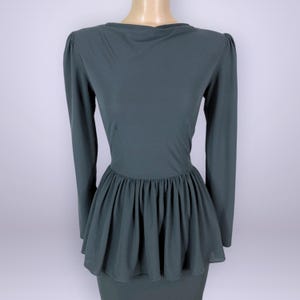 Puede incluir: Un vestido verde oscuro con mangas largas y cintura peplum. El vestido tiene un escote redondo y un corpi&ntilde;o ajustado que se ensancha en la cintura. El peplum est&aacute; hecho de una tela transparente. El vestido se exhibe en un maniqu&iacute;.