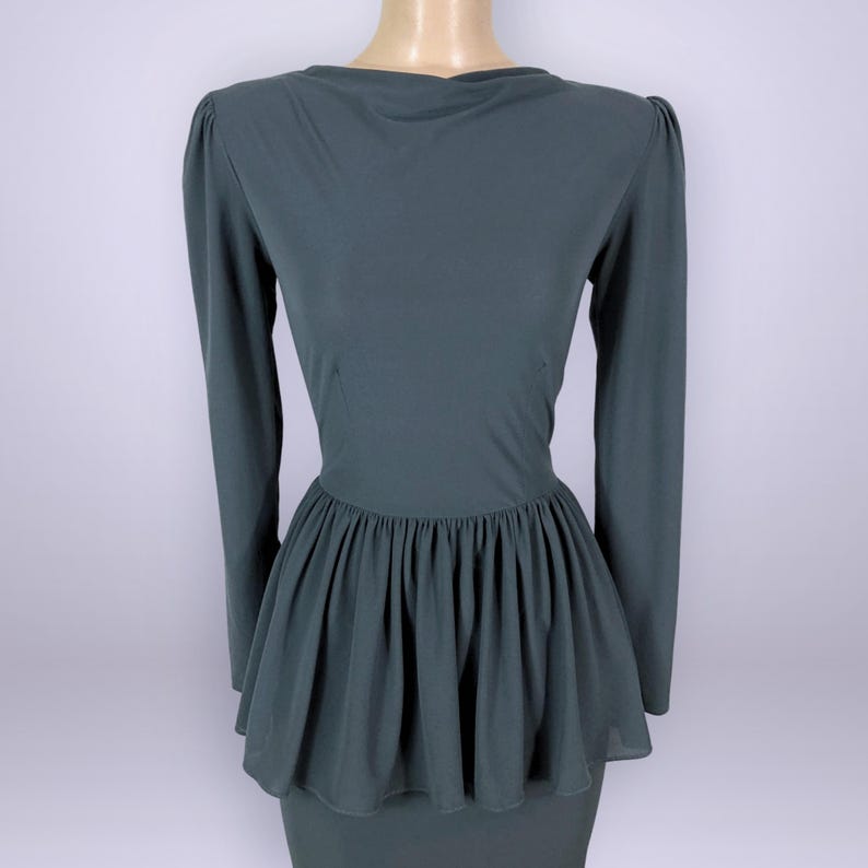 Puede incluir: Un vestido verde oscuro con mangas largas y cintura peplum. El vestido tiene un escote redondo y un corpi&ntilde;o ajustado que se ensancha en la cintura. El peplum est&aacute; hecho de una tela transparente. El vestido se exhibe en un maniqu&iacute;.