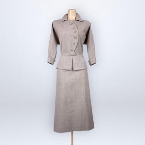 Puede incluir: Un traje vintage de dos piezas en color beige neutro, exhibido en un maniquí. El traje presenta una chaqueta con cuello y botones, con mangas tres cuartos, y una falda a juego hasta la rodilla. Un cinturón ciñe la chaqueta a la cintura.