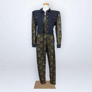 Op de afbeelding: Een vintage zwarte spijkerjas en bijpassende broekset. De jas heeft gouden kanten mouwen en een knoopsluiting. De broek is zwart met een gouden bloemenpatroon. De outfit wordt op een mannequin getoond.