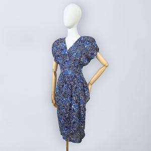 Puede incluir: Un vestido azul vintage con estampado floral. El vestido presenta un escote en V, mangas cortas y cintura entallada. La tela tiene una base azul oscuro con un diseño floral azul claro y blanco. El vestido se exhibe en un maniquí.