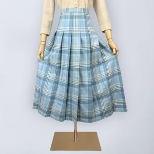 Op de afbeelding: Een lichtblauwe geruite rok met plooien. De rok wordt getoond op een mannequin met een beige top. Het ruitpatroon bevat lichtblauwe, witte en beige lijnen. De rok is knielang.