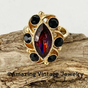 May include: A gold-toned ring featuring a large, marquise-cut, multi-colored gemstone at the center, surrounded by six black, round gemstones. The ring has an ornate, vintage design. The text "Amazing Vintage Jewelry" is at the bottom.