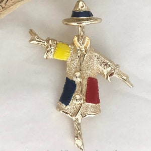May include: A gold-tone metal brooch shaped like a scarecrow wearing a hat with a dark blue band. The scarecrow has a textured coat with a yellow patch on the right sleeve, a dark blue patch on the lower left, and a red patch on the lower right.