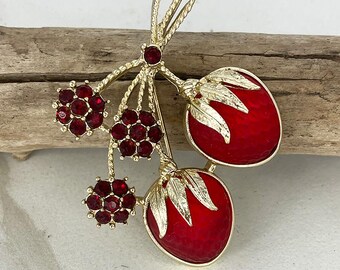 May include: A gold-toned vintage brooch featuring a cluster of red jewel-like berries and two larger red fruit shapes with gold leaf accents. The brooch is set against a wooden background. The text "Amazing Vintage Jewelry" is at the bottom.