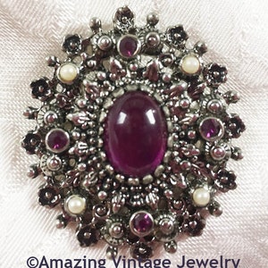 May include: Vintage silver-tone brooch with a central oval, deep purple stone. The brooch features smaller, round purple stones and faux pearls, surrounded by floral and leaf details. The text "Amazing Vintage Jewelry" is at the bottom.