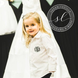 May include: A young girl wearing a white button-down shirt with a monogram on the pocket. The shirt has long sleeves and is tucked into black pants. The girl is standing in front of a white backdrop.