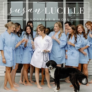 May include: A group of women wearing light blue robes with embroidered initials stand in front of a white house. A black and white dog is in the foreground. The sign above them reads "Susan Lucille Monograms & More".