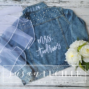 May include: A blue denim jacket with white pearl embellishments on the back. The jacket has white embroidery that says "Mrs. Andrews" and the date "5-11-24". A white veil and a bouquet of white flowers are also in the image.