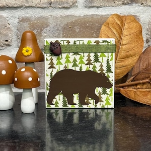May include: A square wooden block featuring a brown bear silhouette against a green forest pattern, topped with a green ribbon and a small acorn. Beside it are two brown mushroom figurines, one with white spots and one with a yellow flower.