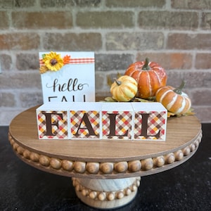 Könnte beinhalten: Ein Holzschild mit dem Text hello FALL und einer Sonnenblume, dazu vier Holzklötze, die das Wort FALL in braunen Buchstaben auf einem rot-orange-gelben Karomuster bilden, sowie drei kleine künstliche Kürbisse.