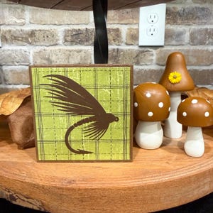 May include: A square wooden sign featuring a dark brown fly fishing lure graphic on a light green plaid background, accompanied by three small decorative mushrooms with brown caps and white spots, one featuring a small yellow flower accent.