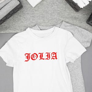 K&ouml;nnte beinhalten: Wei&szlig;es T-Shirt mit dem roten Wort "JOLIA" in einer gotischen Schriftart. Das T-Shirt hat kurze &Auml;rmel und liegt auf einer grauen Oberfl&auml;che. Gefaltete Kleidung befindet sich in K&ouml;rben im Hintergrund.