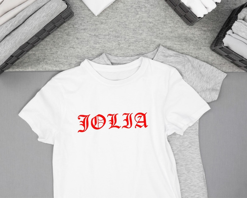 K&ouml;nnte beinhalten: Wei&szlig;es T-Shirt mit dem roten Wort "JOLIA" in einer gotischen Schriftart. Das T-Shirt hat kurze &Auml;rmel und liegt auf einer grauen Oberfl&auml;che. Gefaltete Kleidung befindet sich in K&ouml;rben im Hintergrund.