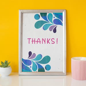 May include: A framed print with the word "THANKS!" in pink. The print has a silver frame and features a teal, blue, and purple abstract design. A pink mug and a small potted succulent are also visible.
