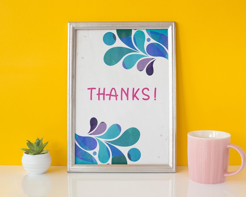 May include: A framed print with the word "THANKS!" in pink. The print has a silver frame and features a teal, blue, and purple abstract design. A pink mug and a small potted succulent are also visible.