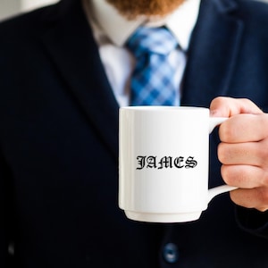K&ouml;nnte beinhalten: Eine wei&szlig;e Keramiktasse mit dem Namen "JAMES" in schwarzer gotischer Schrift. Die Tasse wird von einer Person gehalten, die einen marineblauen Anzug und eine blau gemusterte Krawatte tr&auml;gt. Die Tasse hat eine Standardgr&ouml;&szlig;e.