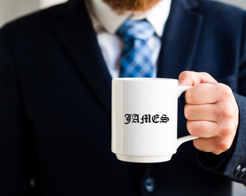 K&ouml;nnte beinhalten: Eine wei&szlig;e Keramiktasse mit dem Namen "JAMES" in schwarzer gotischer Schrift. Die Tasse wird von einer Person gehalten, die einen marineblauen Anzug und eine blau gemusterte Krawatte tr&auml;gt. Die Tasse hat eine Standardgr&ouml;&szlig;e.