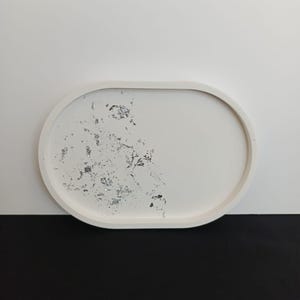 May include: A white, oval-shaped decorative tray with a silver leaf design on one side. The tray has a smooth surface and a raised edge. The background is split between white and black.
