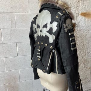 Könnte beinhalten: Dunkelgraue Jacke mit silbernem Totenkopf- und Knochenmuster auf dem Rücken. Die Jacke hat einen pelzgefütterten Kragen, silberne Akzente und mehrere Reißverschlüsse und Knöpfe. Die Jacke wird auf einer Schaufensterpuppe präsentiert.