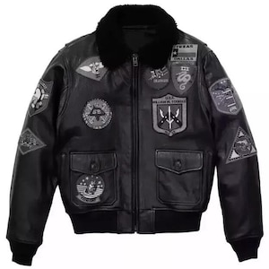 Puede incluir: Chaqueta bomber de cuero negro con cuello de piel sintética negra y múltiples parches bordados. La chaqueta tiene dos bolsillos delanteros y un dobladillo y puños de canalé. Los parches incluyen texto y diseños militares.