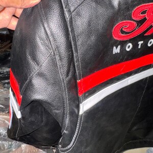 May include: Black leather motorcycle jacket with red and white accents. The jacket features embroidered red lettering that says "Indian Moto". The jacket has a classic design with a padded shoulder area.