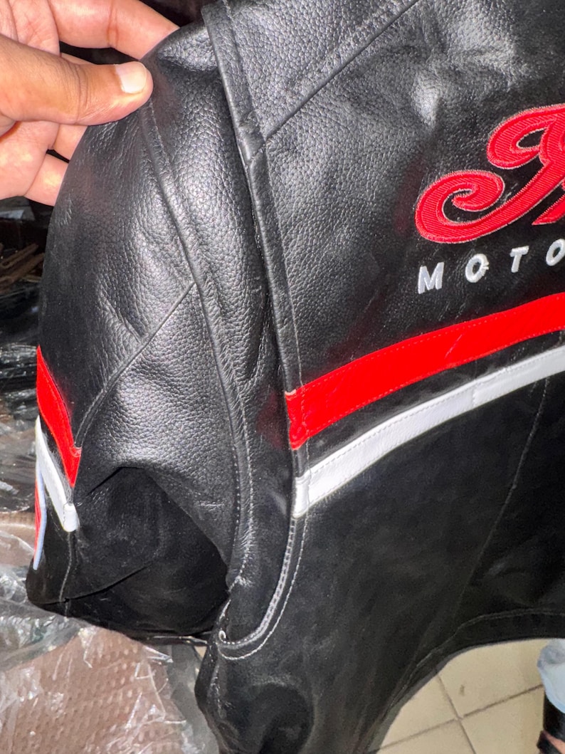 May include: Black leather motorcycle jacket with red and white accents. The jacket features embroidered red lettering that says "Indian Moto". The jacket has a classic design with a padded shoulder area.
